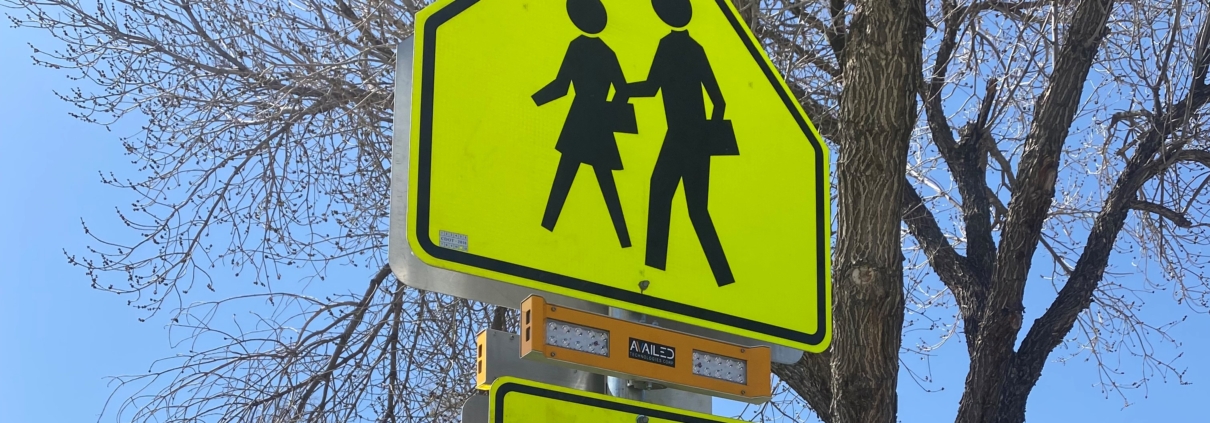 Highlighter yellow Availed RRFB school crossing sign with arrow, with a tree and blue sky in background.